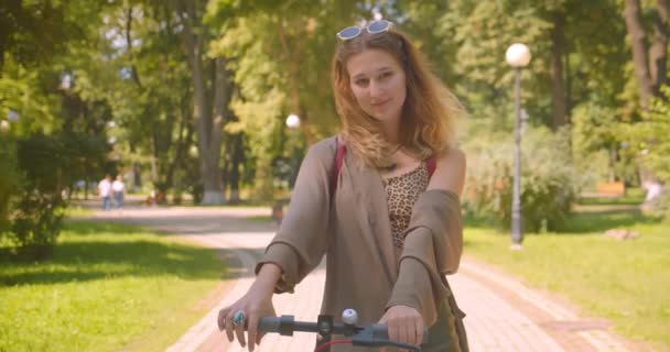 Primo piano ritratto di giovane bella femmina caucasica in piedi con scooter calcio guardando la fotocamera sorridente felicemente nel parco all'aperto — Video Stock