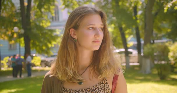 Крупный план портрета молодой белой девушки, счастливо улыбающейся, стоя в парке на открытом воздухе — стоковое видео