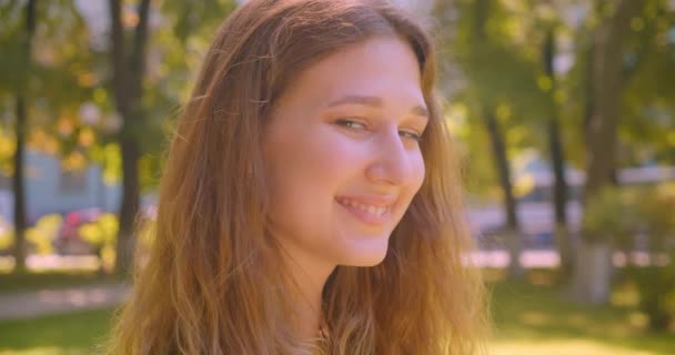 Närbild porträtt av unga söta kaukasiska kvinna tittar på kamera leende glatt poserar stående i parken utomhus — Stockvideo