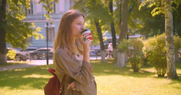 Nahaufnahme Seitenansicht Porträt der jungen niedlichen kaukasischen Frau, die in die Kamera schaut, Kaffee trinkt, glücklich lächelnd im Park im Freien steht — Stockvideo