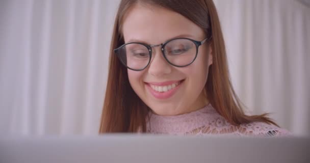 Крупный план портрета молодой привлекательной кавказской предпринимательницы в очках, печатающей на ноутбуке, радостно улыбающейся сидя на диване в помещении — стоковое видео