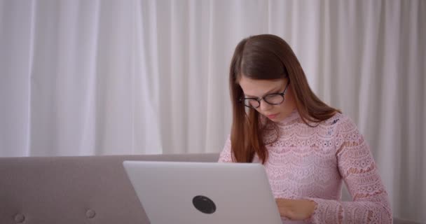 Крупный план портрета молодой привлекательной кавказской предпринимательницы в очках, печатающей на ноутбуке, смотрящей в камеру и радостно улыбающейся сидящей на диване в помещении — стоковое видео
