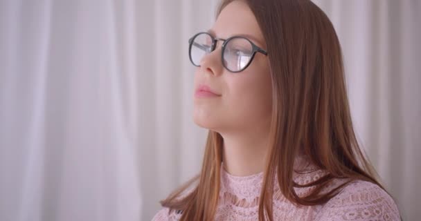 Nahaufnahme Porträt einer jungen attraktiven kaukasischen Geschäftsfrau mit Brille, die glücklich lächelnd in die Kamera schaut, während sie drinnen auf der Couch sitzt — Stockvideo