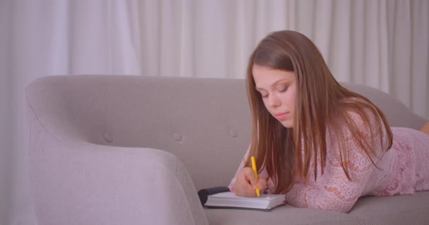 Nahaufnahme Porträt einer jungen attraktiven kaukasischen Frau, die in Tagebuch schreibt und auf der Couch in der Wohnung liegt — Stockvideo