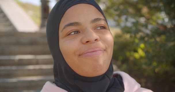 Крупним планом портрет молодої привабливої афроамериканської мусульманської дівчини в хіджабі, дивлячись на камеру, яка щасливо посміхається в міському місті на відкритому повітрі — стокове відео