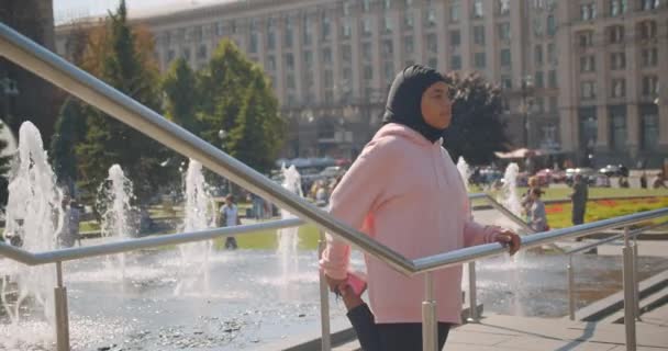 Closeup portrait of young attractive african american muslim girl in hijab stretching next to fountains in urban city outdoors — Stock Video