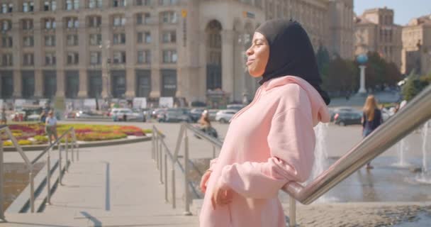 Primer plano retrato de la joven atractiva chica musulmana afroamericana en hijab de pie junto a fuentes sonriendo felizmente en la ciudad urbana al aire libre — Vídeo de stock