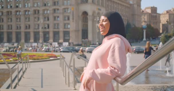 Closeup portrait of young attractive african american muslim girl in hijab standing next to fountains smiling cheerfully in urban city outdoors — Stock Video