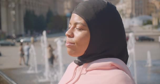 Closeup portrait of young attractive african american muslim girl in hijab standing next to fountains smiling cheerfully in city outdoors — Stock Video