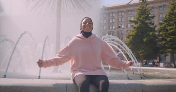 Gros plan portrait de jeune fille musulmane afro-américaine attrayante en hijab souriant joyeusement assis avec des fontaines en arrière-plan dans la ville urbaine en plein air — Video