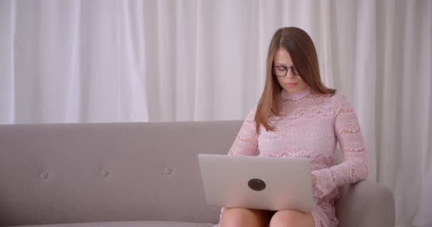 Крупный план портрета молодой привлекательной белой женщины с помощью ноутбука, смотрящей в камеру и счастливо улыбающейся сидящей на диване в помещении — стоковое видео