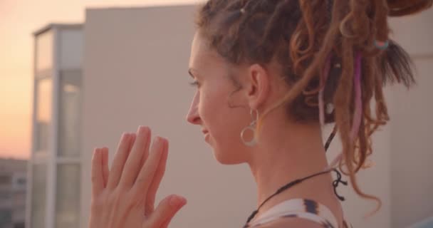 Gros portrait de femme spirituelle avec dreadlocks faire du yoga méditant sur le toit avec beau coucher de soleil paysage de la ville sur le fond — Video