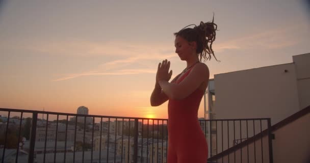 Vista lateral close-up retrato de mulher espiritual com dreadlocks praticando ioga sorrindo alegremente no telhado com belo pôr do sol no fundo — Vídeo de Stock