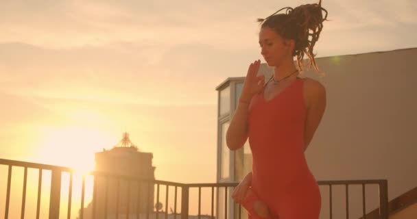Retrato de close-up de fêmea hipster com dreadlocks praticando asanas de ioga que se estendem no telhado com belo pôr do sol paisagem da cidade no fundo — Vídeo de Stock