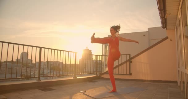 Retrato de cerca de una mujer consciente con rastas practicando yoga que se extiende en la azotea con hermoso paisaje de la ciudad puesta de sol en el fondo — Vídeos de Stock