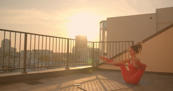 Närbild porträtt av unga sportig hona med dreadlocks öva yoga asanas på taket i staden med vacker soluppgång på bakgrunden — Stockvideo