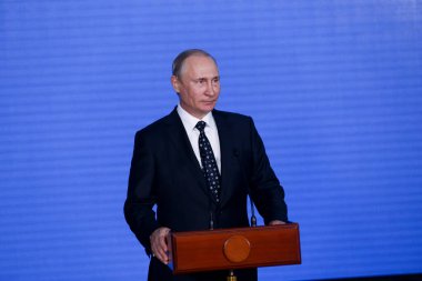 Rusya Federasyonu Başkanı Vladimir Putin kürsünün arkasında duruyor ve mavi bir duvarın arkasından konuşuyor.