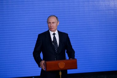 Rusya Federasyonu Başkanı Vladimir Putin kürsünün arkasında duruyor ve mavi bir duvarın arkasından konuşuyor.