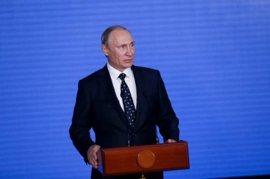 Rusya Federasyonu Başkanı Vladimir Putin kürsünün arkasında duruyor ve mavi bir duvarın arkasından konuşuyor.