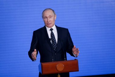 Rusya Federasyonu Başkanı Vladimir Putin kürsünün arkasında duruyor ve mavi bir duvarın arkasından konuşuyor.