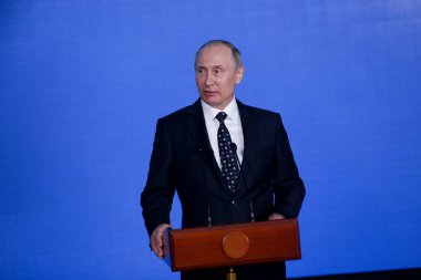 Rusya Federasyonu Başkanı Vladimir Putin kürsünün arkasında duruyor ve mavi bir duvarın arkasından konuşuyor.