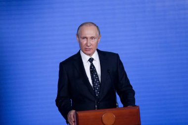 Rusya Federasyonu Başkanı Vladimir Putin kürsünün arkasında duruyor ve mavi bir duvarın arkasından konuşuyor.