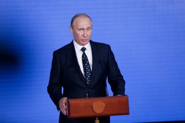 Rusya Federasyonu Başkanı Vladimir Putin kürsünün arkasında duruyor ve mavi bir duvarın arkasından konuşuyor.