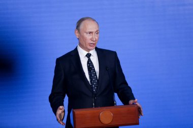Rusya Federasyonu Başkanı Vladimir Putin kürsünün arkasında duruyor ve mavi bir duvarın arkasından konuşuyor.