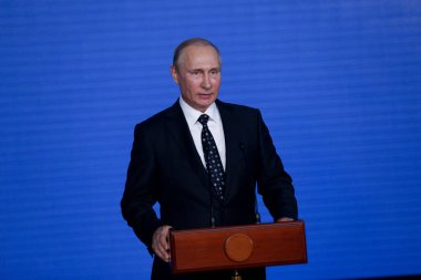 Rusya Federasyonu Başkanı Vladimir Putin kürsünün arkasında duruyor ve mavi bir duvarın arkasından konuşuyor.