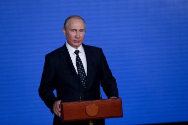 Rusya Federasyonu Başkanı Vladimir Putin kürsünün arkasında duruyor ve mavi bir duvarın arkasından konuşuyor.