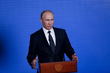 Rusya Federasyonu Başkanı Vladimir Putin kürsünün arkasında duruyor ve mavi bir duvarın arkasından konuşuyor.