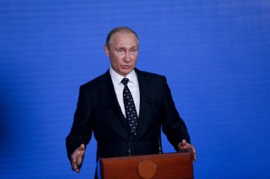 Rusya Federasyonu Başkanı Vladimir Putin kürsünün arkasında duruyor ve mavi bir duvarın arkasından konuşuyor.