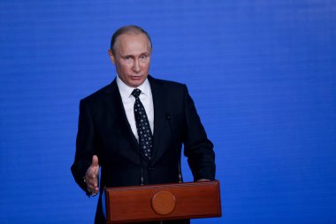 Rusya Federasyonu Başkanı Vladimir Putin kürsünün arkasında duruyor ve mavi bir duvarın arkasından konuşuyor.