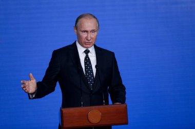 Rusya Federasyonu Başkanı Vladimir Putin kürsünün arkasında duruyor ve mavi bir duvarın arkasından konuşuyor.