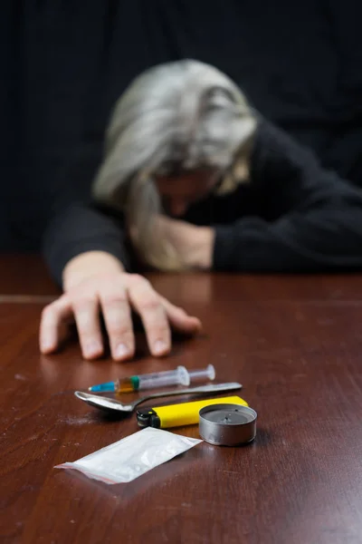 Narkoman u stolu táhne ruku do stříkačky s dávkou. Koncepce proti drogám. — Stock fotografie