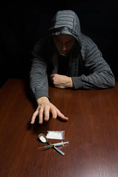 Narkoman u stolu táhne ruku do stříkačky s dávkou. Koncepce proti drogám. — Stock fotografie