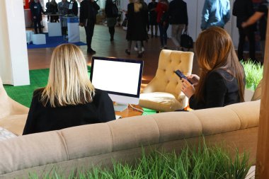 İki iş kadını beyaz ekranlı bir bilgisayarda kanepede oturuyor. İnsanların siluetleri etrafta dolaşıyor. Mocap.                       