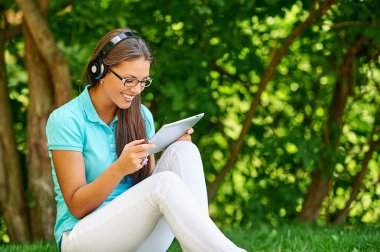 Kulaklıklı, müzik dinleyen ve yeşil çimlerde otururken tablet kullanan mutlu bir kadın.