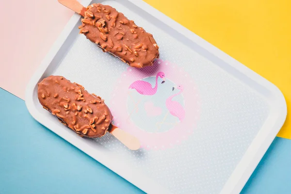 Dessert chocolate ice cream bars with nuts, wooden stick on blue and yellow pastel background. Eskimo in blue vintage tray with pink flamingosand dots. Summer temptation. Indoors closeup. — Stock Photo, Image