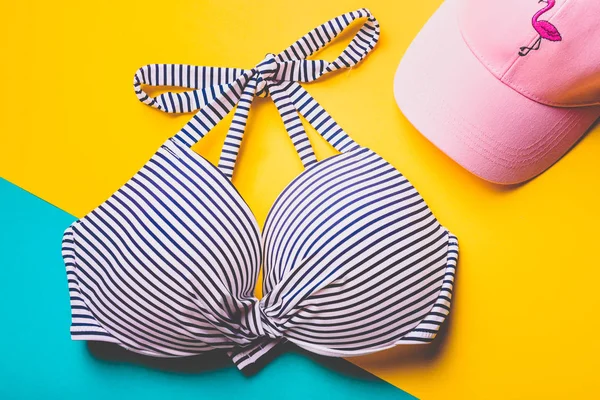 Vue du dessus du soutien-gorge de bain rayé bleu et blanc et chapeau de plage rose avec flamant rose sur fond pastel bleu et jaune. Espace de copie. Concept de vacances . — Photo