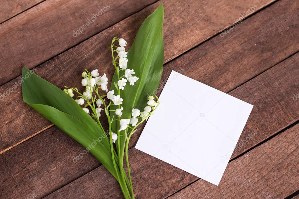 delicate white lilies of the valley with green leaves on a brown wooden background with a sheet of white paper for text