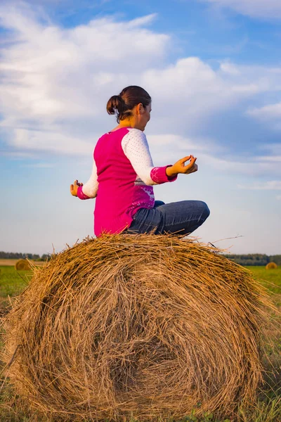 Женщина медитирует в поле против голубого неба на стоге сена на закате — стоковое фото