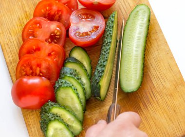 Bıçaklı bir el, dilimlenmiş domateslerin bulunduğu kesme tahtasındaki salatalığı keser..