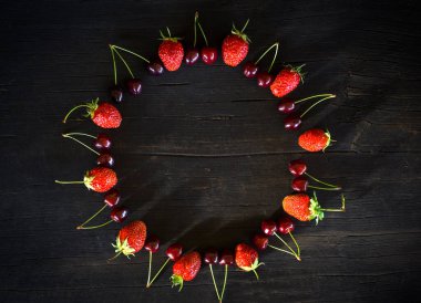 Çilekler ve kirazlar koyu ahşap bir zemin üzerinde uzayın bir kopyasıyla daire şeklinde serilmiştir..