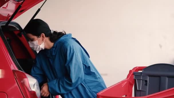 Ung kvinde bærer medicinsk ansigtsmaske mod corona virus covid-19 i parkeringskælderen. Kunde lastning bil med mad på supermarked parkeringsplads. Fødevaremangel. Panikkøb og hamstring. – Stock-video