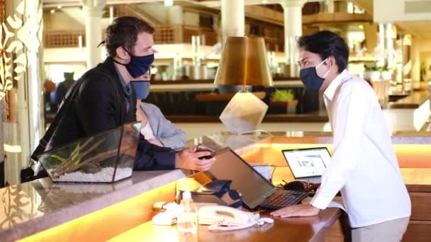 Paar en receptioniste aan de balie in het hotel dragen medische maskers als voorzorgsmaatregel tegen het virus. Paar op een zakenreis doen check-in in het hotel — Stockvideo