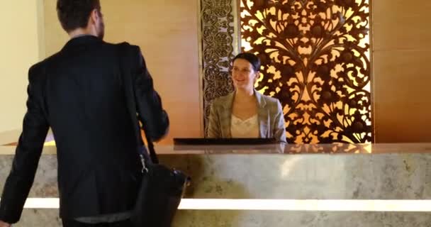 ビジネスマンがチェックインのためにホテルのロビーに入る。カウンター受付で若いビジネスマンや受付嬢 — ストック動画