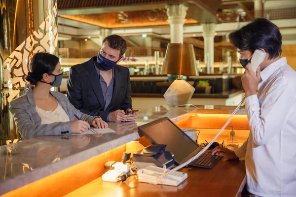 Пара и секретарша в отеле в медицинских масках в качестве меры предосторожности против вируса. Пара в командировке, регистрирующаяся в отеле — стоковое фото
