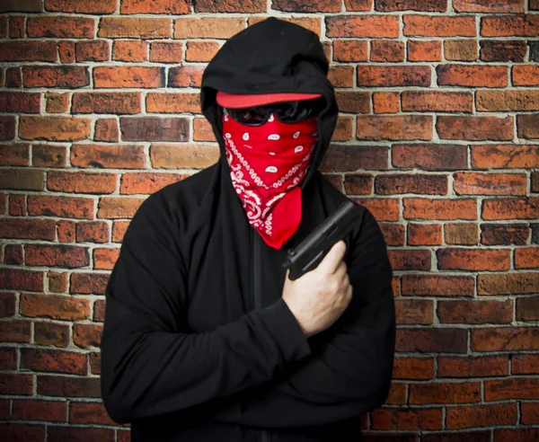 Hidden man with hood, scarf and gun in front of a brick wall — Stock Photo, Image