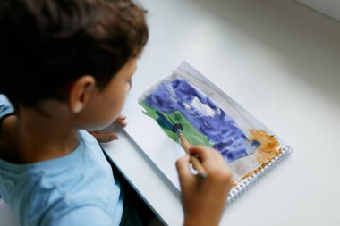 Mavi tişörtlü kıvırcık saçlı çocuk beyaz bir masada otururken boyalı bir resim çiziyor.